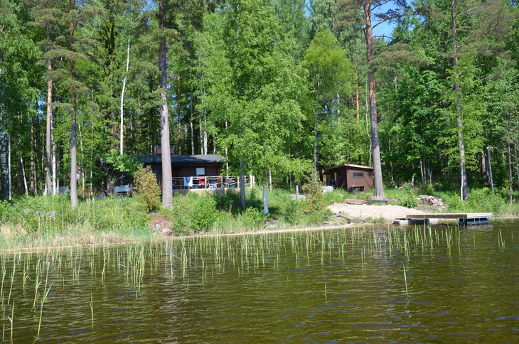 Näkymä järveltä rantaan, jossa puiden katveessa näkyy kaksi mökkirakennusta ja pieni laituri.