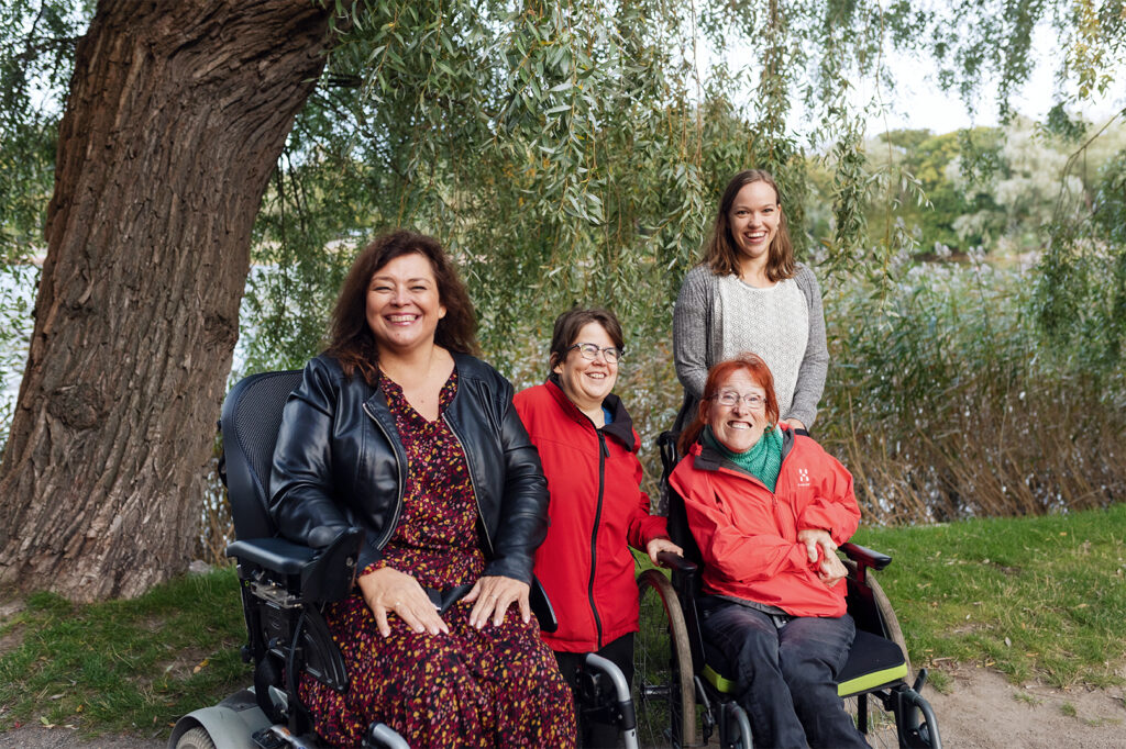 Neljä naista ulkona hopeapajun katveessa. Yksi naisista istuu sähköpyörätuolissa, toinen manuaalipyörätuolissa. Toiset kaksi seisovat. Kaikki hymyilevät.