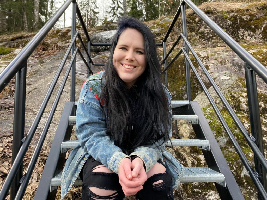 Emma Sofianna Söderholm istuu metallisilla rappusilla kallioisessa maisemassa. Söderholm katsoo hymyillen kameraan. Hänellä on pitkä musta tukka, farkkutakki ja mustat farkut, jotka on polvesta rikki.