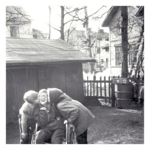 Mustavalkoinen vanha kuva. Matalan puurakennuksen edessä pyörätuolissa istuva silmälasipäinen lapsi (Lissu Lundström), jonka molemmilla puolilla tytöt kumartuvat antamaan hänelle suukkoa poskille.