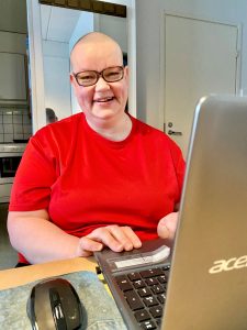 Punaiseen t-paitan pukeutunut siilitukkainen silmälasipäinen Jaana Huhtala istuu kannettavan tietokoneen ääressä ja hymyilee.