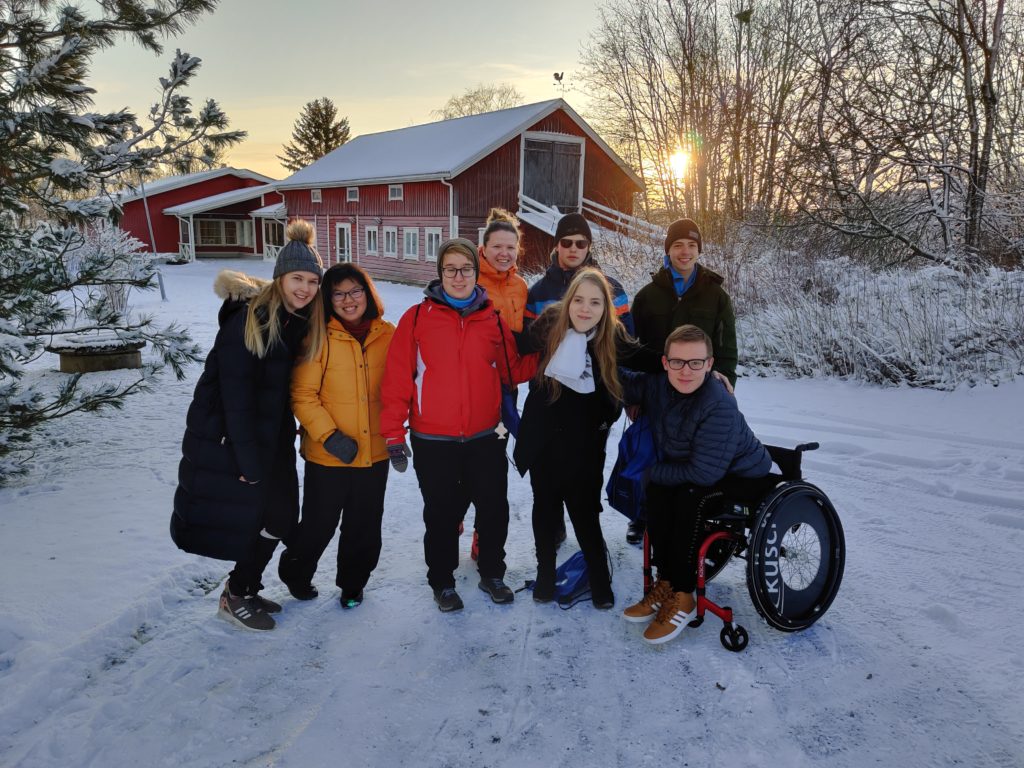 Kahdeksan nuorta aikuista ryhmässä ulkona talvisessa maisemassa. Tuastalla lumen peitätmiä puita ja pensaita ja punainen hirsinen rakennus. Seitsemän nuorista seisoo, yksi istuu pyörätuolissa.