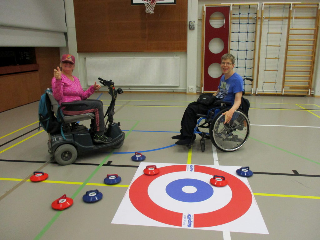 Kaksi pelaajaa sähkömopossa ja pyörätuolissa ihastelevat lattiacurlingin lopputilannetta liikuntasalissa. Pelaajat katsovat kameraan ja hymyilevät leveästi.
