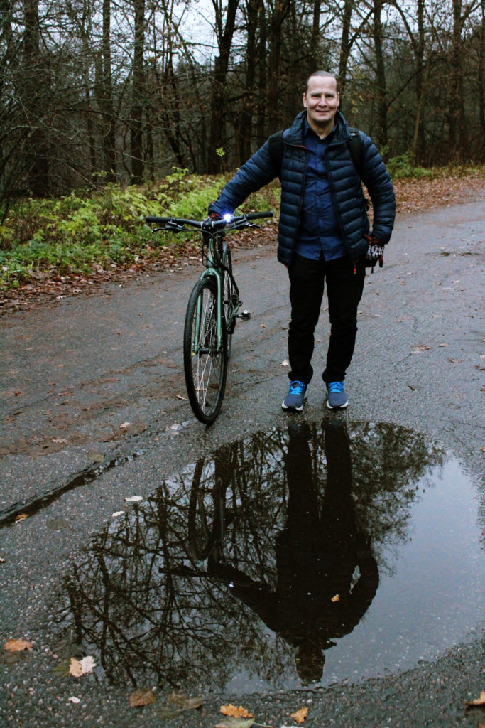 Kari Toiviainen seisoo ulkona asfalttitiellä pyöränsä vieressä. Edessä on vesilätäkkö, johon Karin kuva heijastuu. On syksy, lehdet ovat tippuneet puista ja taivas on harmaa.
