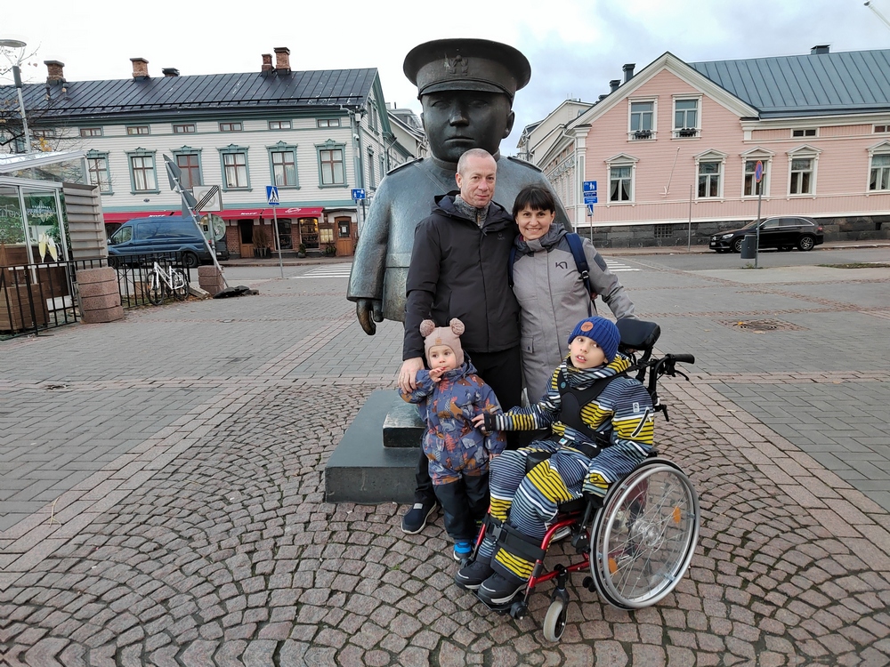 Kuvassa Oulun toripoliisi-patsaan edessä otettu kuva, jossa takana seisovat mies ja nainen ja edessä poika istuu pyörätuolissa ja vieressä seisoo pienempi poika. 