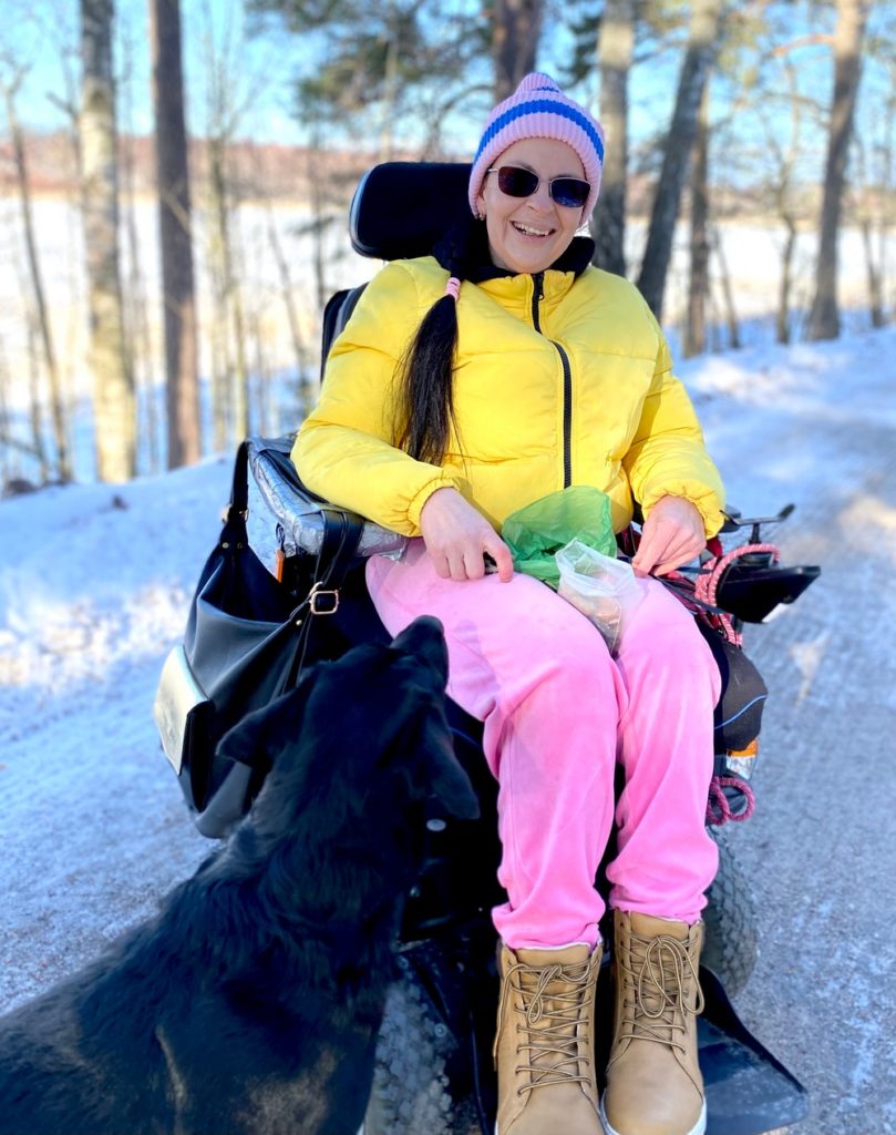 Lumisessa ja aurinkoisessa säässä nainen istuu sähköpyörätuolissa ja katsoo hymyillen kameraan. Edessä vasemmalla musta koira katsoo naista.