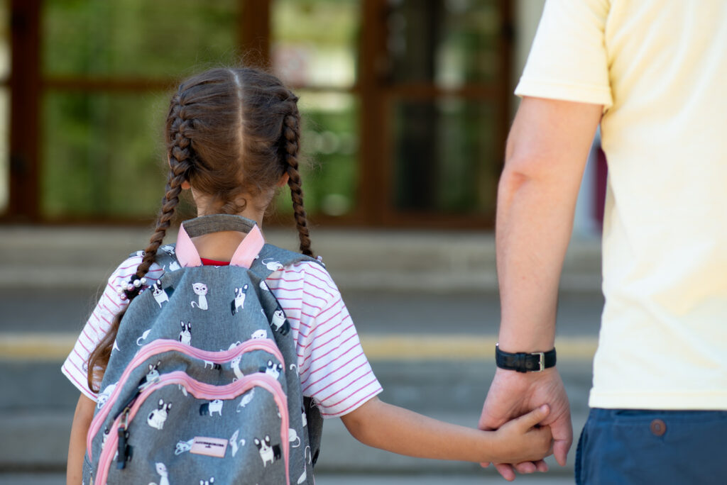 Lapsi, jolla on selässään koulureppu, pitää kiinni aikuisen miehen kädestä.