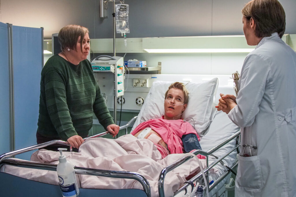 Keskellä kuvaa henkilö makaa sairaalavuoteessa ja katsoo kohti lääkäriä. Vasemmalla kuvassa kolmas henkilö seisoo ja nojaa sairaalasänkyyn katse lääkärissä. 