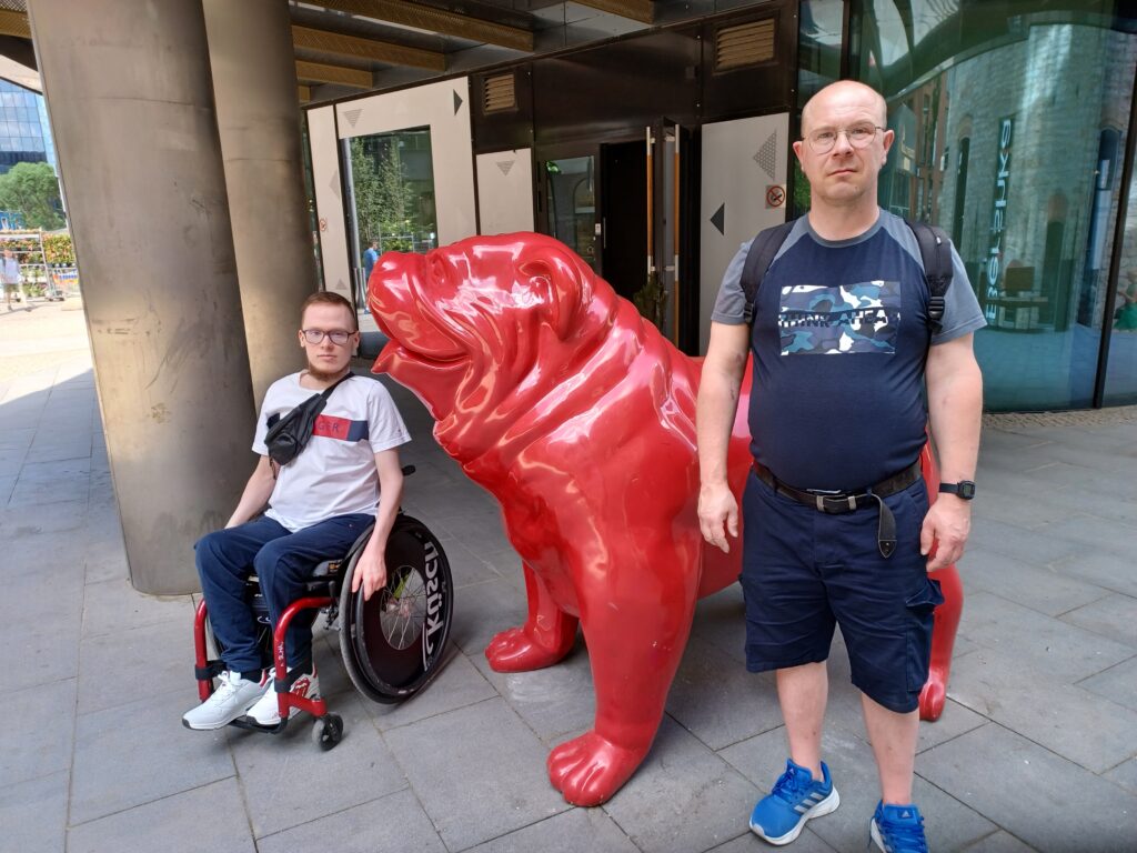 Yksi mies pyörätuolissa ja toinen seisoo, välissä keskellä punainen iso koirapatsas.