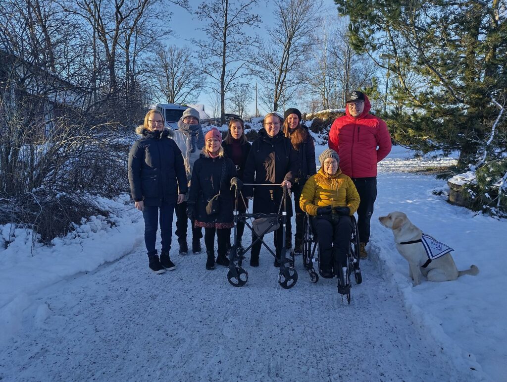 Kahdeksan ihmistä kahdessa rivissä ulkovaatteissa. Maa on peittynyt lumaan ja aurinko paistaa. 