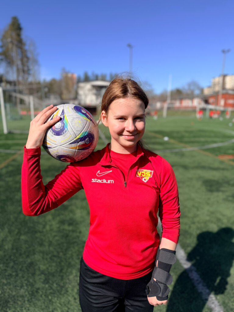 Matilda Laurell seisoo jalkapallokentällä punaisessa Vantaan Jalkapalloseuran paidassa ja pitelee jalkapalloa oikean olkapäänsä päällä. Hän katsoo hymyillen kameraan.