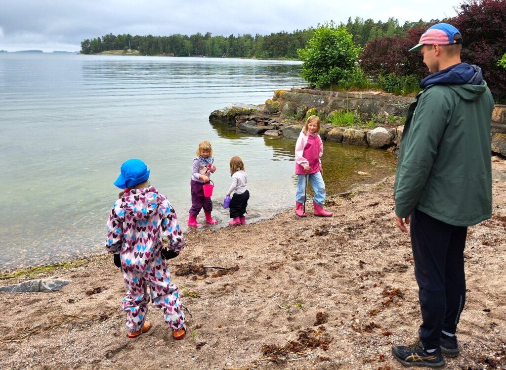Rannalla vesirajassa leikkii neljä lasta värikkäissä vaatteissa ja kumisaappaat jalassa. Yhdellä lapsista on ämpäri kädessä. Oikealla seisoo mies lippalakki päässä ja katselee rannalle.