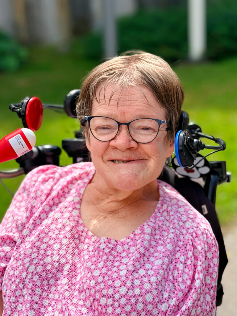 Eeva Kinnunen istuu sähköpyörätuolissaan ja hymyilee. Eevalla on lyhyet ruskeat hiukset, silmälasit ja vaaleanpunainen paita, jossa on valkoisia kukkia.