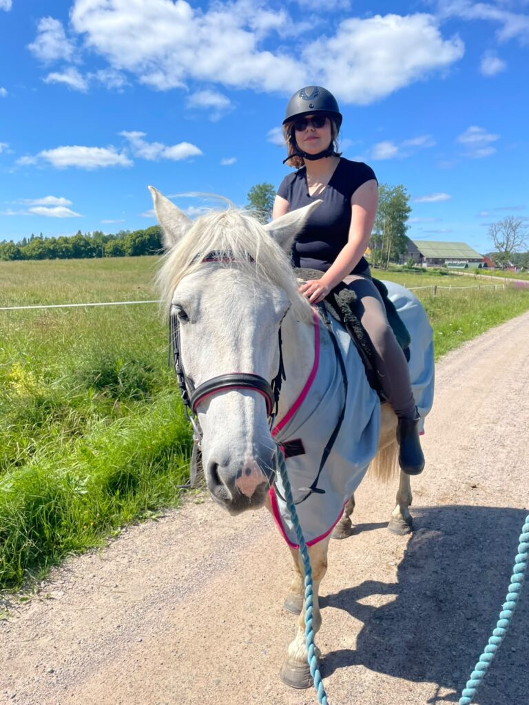 Nella Nieminen ratsastaa valkoisella hevosella kesäisen niityn vieressä. Nellalla on musta kypärä ja musta t-paita.