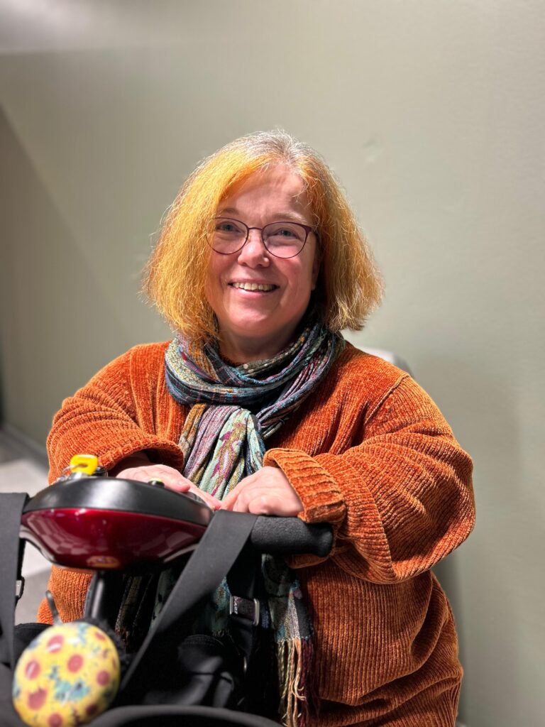 Paula Ahti. Paulalla on punertavat lyhyet hiukset, silmälasit ja oranssi neule Hän nojaa sähkömopoon.